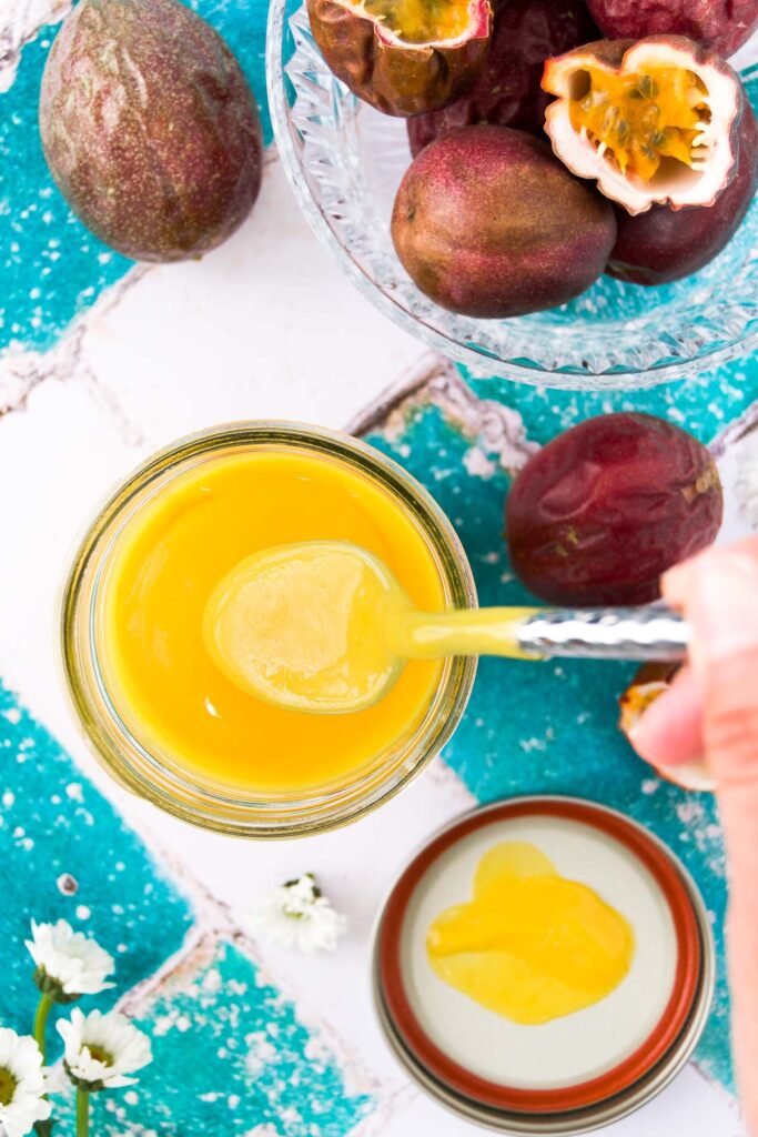 a spoon dips into a jar of passionfruit curd on a teal and white tiled surface