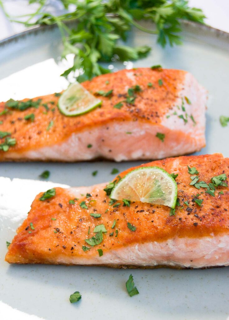 two pieces of seared salmon on a white plate garnished with fresh herbs and lemon